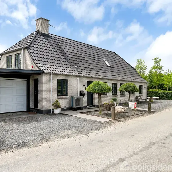 Et hvidt hus med skråt tag står ved en vej. Huset har en garageport og er omgivet af grus og velplejede træer. Solrig dag og grøn vegetation omkranser området.