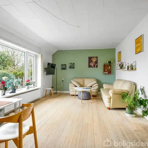 Stue med lyse trægulve, beige sofaer og grøn væg. Store vinduer til venstre med udsigt til have. Bord med stole i forgrunden, planter og kunst på væggene.