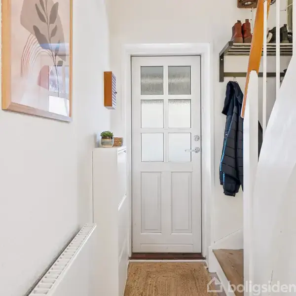 Dør med glasruder står lukket i en entré. Til højre hænger frakker på en stumtjener. Venstre væg har billeder og en hylde med en potteplante.