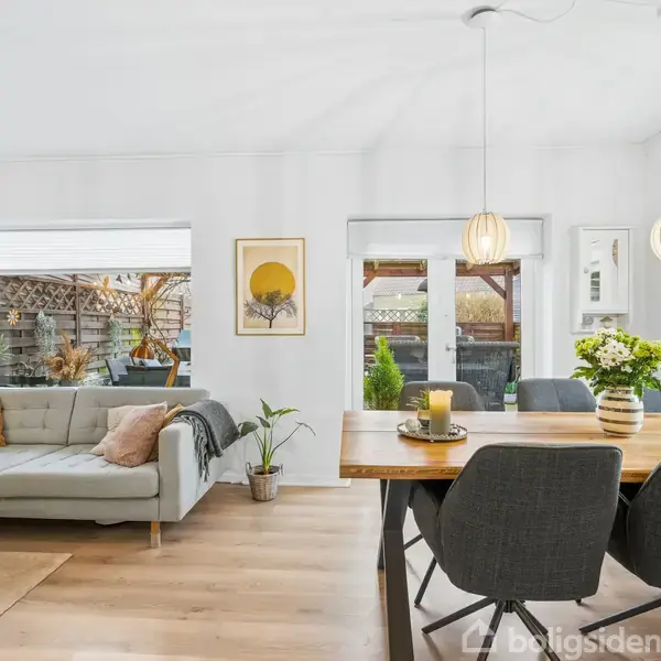 En moderne stue indeholder en grå sofa og et spisebord i træ omgivet af stole. Rummet er lyst med planter, lysdekorationer og kunst på væggene samt udsigt til en terrasse.