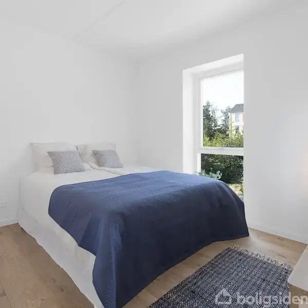 En seng med hvidt sengetøj og et blåt tæppe står i et lyst, minimalistisk soveværelse med trægulv. Et lille bord med lampe ses ved vinduet, som viser træer udenfor.