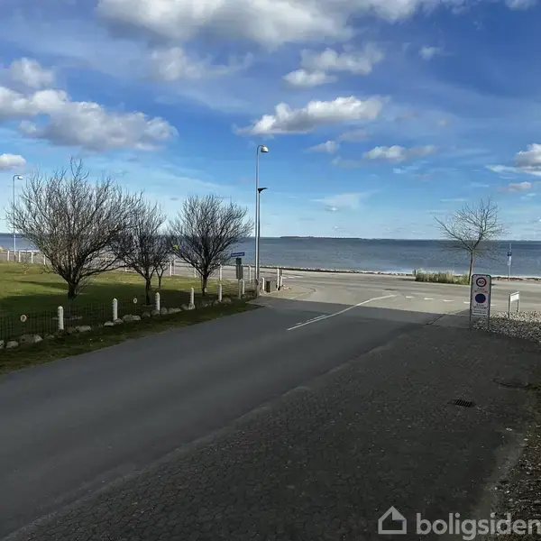 Vejen strækker sig mod havet, med træer langs venstre side. I baggrunden skimtes en strand og blå himmel med skyer. Der ses ingen tekst i billedet.
