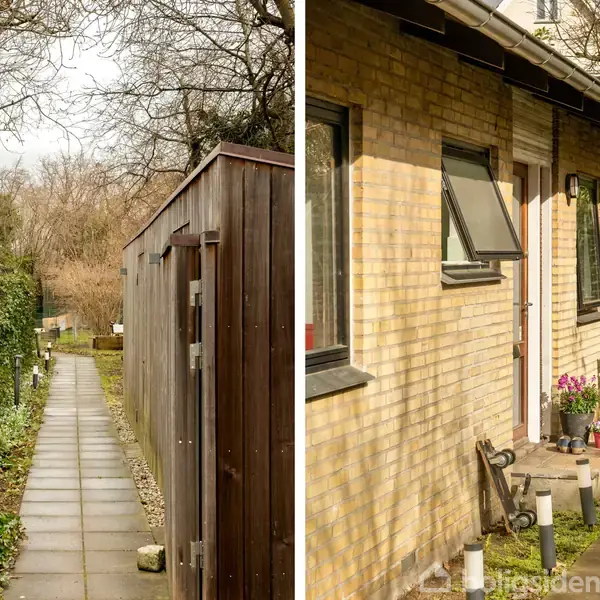 Smalt havegang med flisebelægning ved siden af en træbeklædt hegn. En murstensbygning med åbent vindue og blomster i potter står ved siden af en bænk og planter.