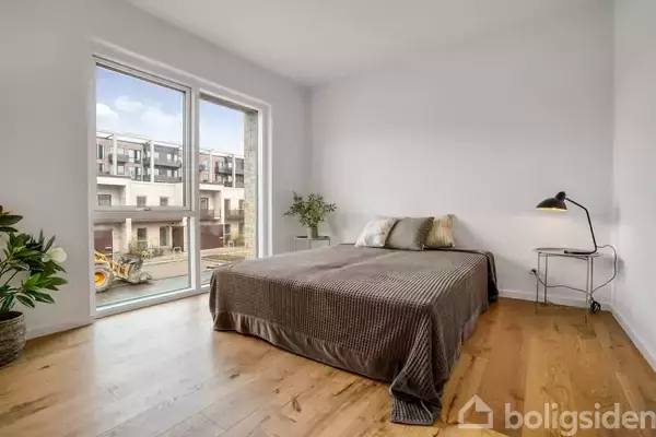 En seng står i et lyst rum med trægulv og en stor vindue, der viser en byggeplads udenfor. Ved siden af sengen er der en minimalistisk lampe og et lille bord.