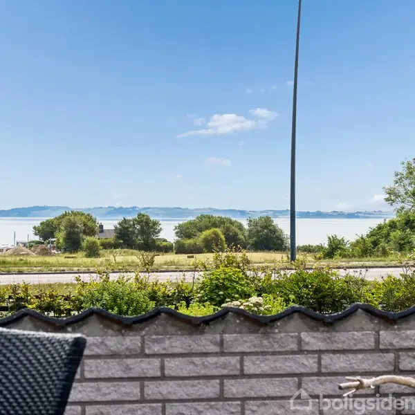 En stol står på en terrasse med udsigt over en vej og frodig vegetation mod en blå sø eller hav med træer i baggrunden under en klar, blå himmel.