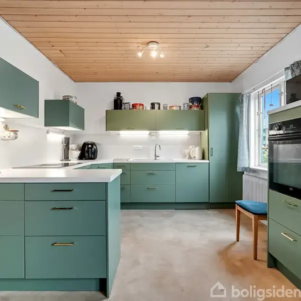 Et moderne køkken med grønne skabe og skuffer, hvor der ses en ovn indbygget i et skab. Køkkenmaskiner står på køkkenbordet, og en træstol står ved vinduet.