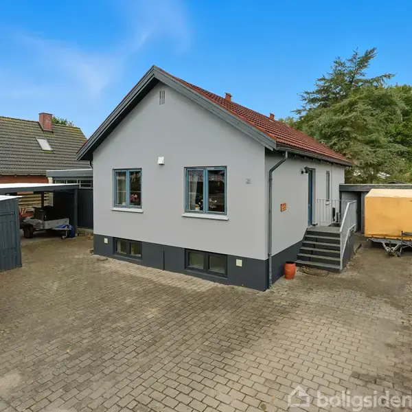 Gråt hus med rødt tag står på brolagt indkørsel. Ved siden af huset er en carport og trailer. Omgivet af grønne træer og nabolagets huse. Nummer 28 på postkassen.