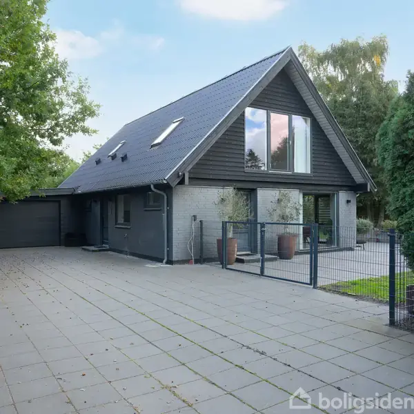 Et moderne, gråt hus med skråt tag og store vinduer står på en flisebelagt indkørsel. Det omkranses af træer og hegn i en rolig boligområde.