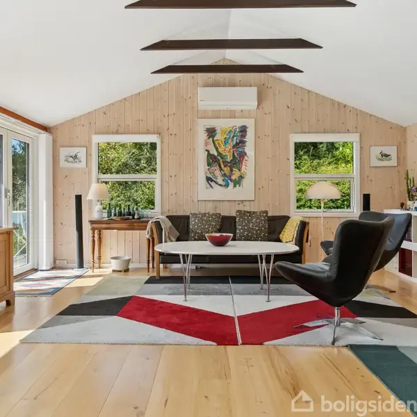 Stue med møbler: en sofa, lænestol, og sofabord på et farverigt tæppe. Trævægge med kunstværker og store vinduer, der giver udsigt til grønne omgivelser. Fjernsyn på skænk til venstre.