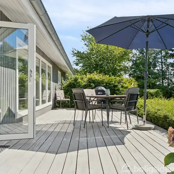 En træterrasse med et sæt havemøbler og stor parasol. Terrassen har en åben dør til huset og omgives af grønne buske og træer i baggrunden.