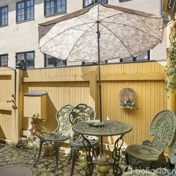 En grønmetal bord- og stolgruppe står på en brostensbelagt gårdhave under en stor, mønstret parasol. Væggen i baggrunden er malet gul og dekoreret med plantekasser.