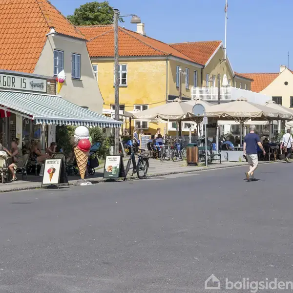 En mand går hen ad en solrig gade med caféer og cykler i forgrunden. Folk sidder ved borde under parasoller. Skiltet siger "DRAGØR IS". En stor isvaffel står ved siden af.