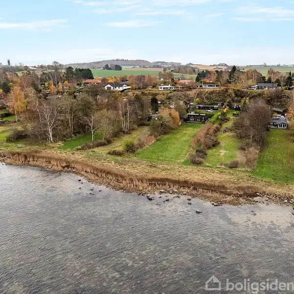 Kyststrækning med græs og træer strækker sig ind i baggrunden, hvor huse er omgivet af efterårsfarvede træer. Der er vand i forgrunden og åbent land i horisonten.