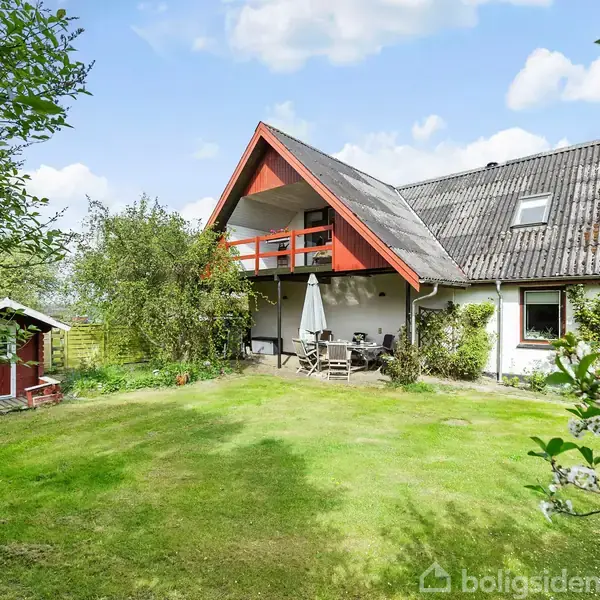 Hus med rødt tag og balkon, omgivet af grøn have med træer og græs. En parasol står ved et bord med stole i haven.