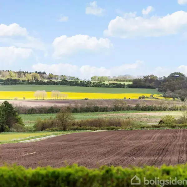 Markeret jord ligger i forgrunden, omgivet af åbne marker med gule afgrøder og skovklædte bakker i baggrunden under en blå himmel med lette skyer.