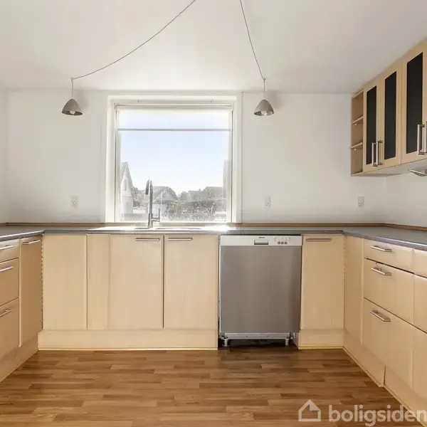 Køkken med lyst træinventar. Opvaskemaskine under vindue, overskabe med glaslåger til højre. Rummelige bordplader omkranser, lys strømmer ind. Trælaminatgulv og hvide vægge kompletterer det enkle design.