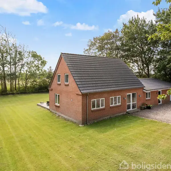 En rødstensvilla med sort tegltag står stille på en velplejet græsplæne, omgivet af træer og buske. Der ses en flisebelagt terrasse og en grusbelagt indkørsel.