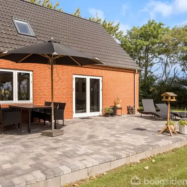 Murstenshus med terrasse og have. På terrassen står et sort parasol over et bord- og stolesæt. Liggestole og fuglehus ses også. Omkranset af græsplæne og træer.