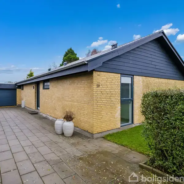 Et murstenshus står ved en flisebelagt indkørsel, omgivet af en grøn hæk og træer i baggrunden, under en blå himmel med skyer. Nummeret "59" ses ved døren.