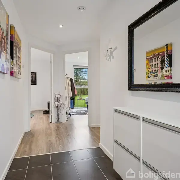 Spejl i entré reflekterer farverige malerier. Lang gang med sort flisegulv og hvid kommode fører til rum med trægulv. Vindue i baggrunden viser en udendørs have.