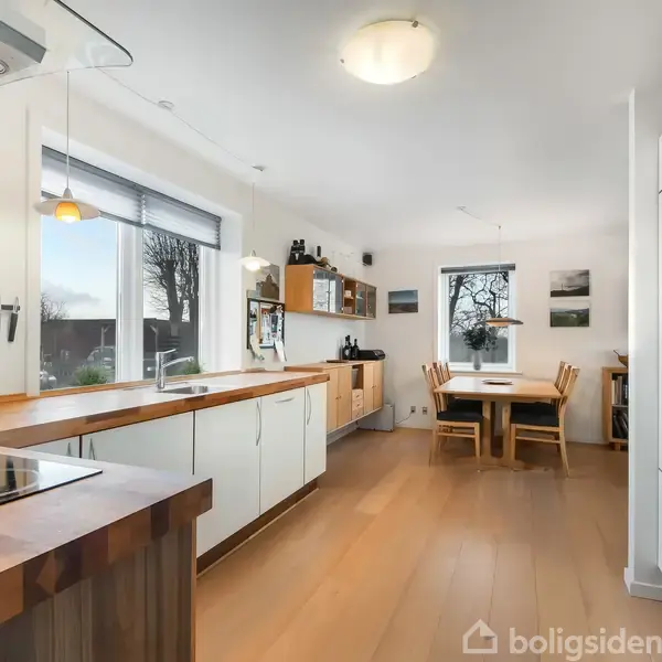 Køkken med hvide skabe og træbordplader strækker sig mod spiseområde i lys, rummelig indretning; store vinduer kaster naturligt lys over rummet, hvor et spisebord har fire stole.