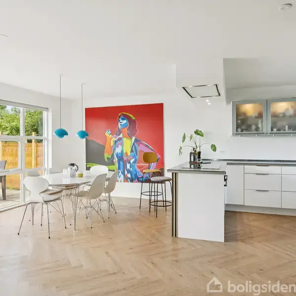 Køkken-alrum med åbne glasdøre mod terrasse. Hvidt køkken med ø, spisebord og stole. Vægkunst med en farverig figur i baggrunden. Trægulve og moderne indretning dominerer rummet.
