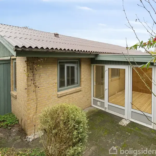Et lille murstenshus med store vinduespartier står stille i en have omgivet af buske og træer. En fliseterrasse ligger foran huset.