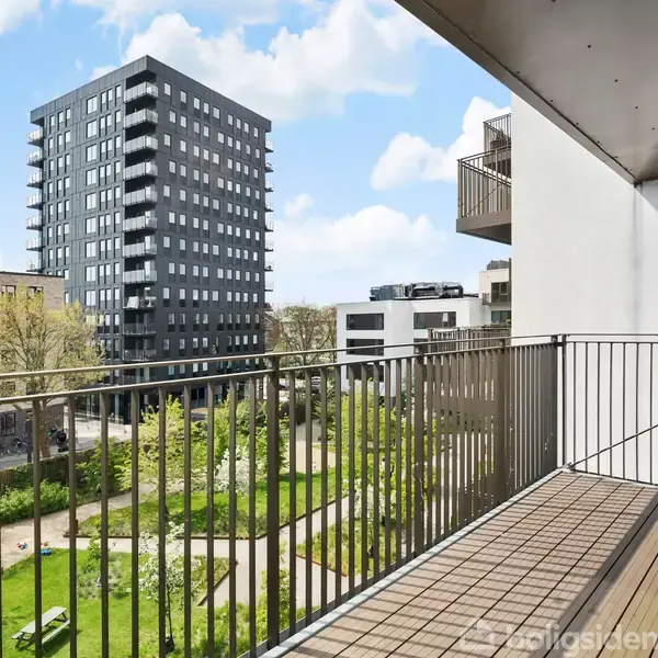 En balkon med metalrækværk strækker sig over en lejlighedsbygning. Udsigt over grøn gårdhave med græsplæne og blomsterbede. Moderne højhuse i baggrunden under en blå himmel.