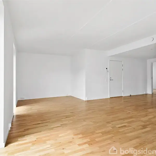 Stue med træparketgulv uden møbler, gennemstrømmet af lys fra store vinduer. Hvide vægge og lofter skaber en lys og neutral atmosfære i rummet.