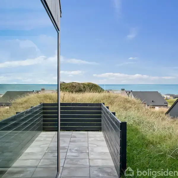 Terrasse med glasværn reflekterer himlen; beliggende ved græsbevokset klitlandskab. I baggrunden ses sommerhuse med sorte tage og det fjerne hav under en blå, skyet himmel.