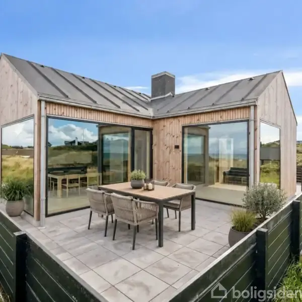 Et moderne sommerhus med store vinduer og træbeklædning står på en flisebelagt terrasse med spisebord og stole. Det er omgivet af græsarealer og har udsigt til et åbent landskab.