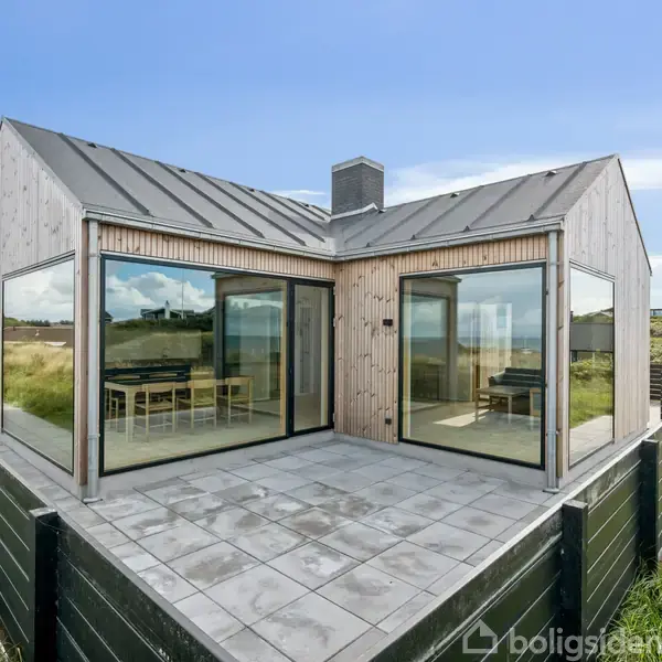Et moderne sommerhus med store glaspartier står på en flisebelagt terrasse. Det ligger i et grønt naturlandskab med blåt himmel som baggrund.