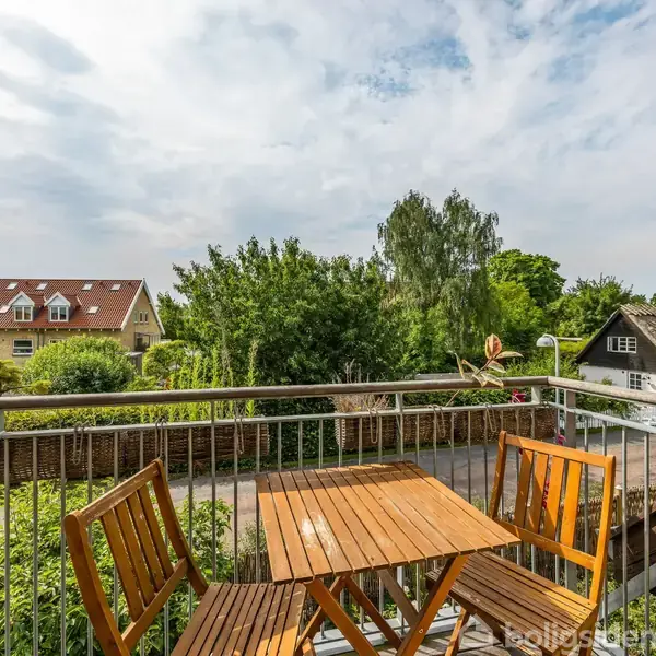 Et sæt træmøbler står stille på en altan med udsigt over en grøn have og boligområde. Skov, huse og en delvis skyet himmel præger omgivelserne.
