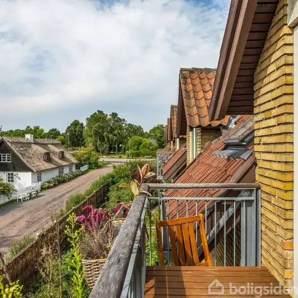 Altan med træstol i et boligområde med rækkehusfacader og røde tage. Udsigt over en lille vej med et traditionelt hus med stråtag omgivet af grønt og træer.