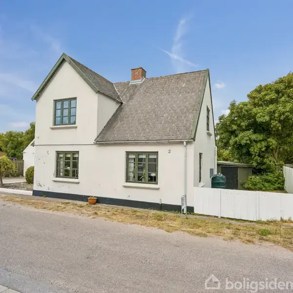 Et hvidt, toetagers hus står på en hjørnegrund ved en rolig vej, omgivet af træer og en lav hvid stakithegn. Husnummeret "2" ses ved indgangen.