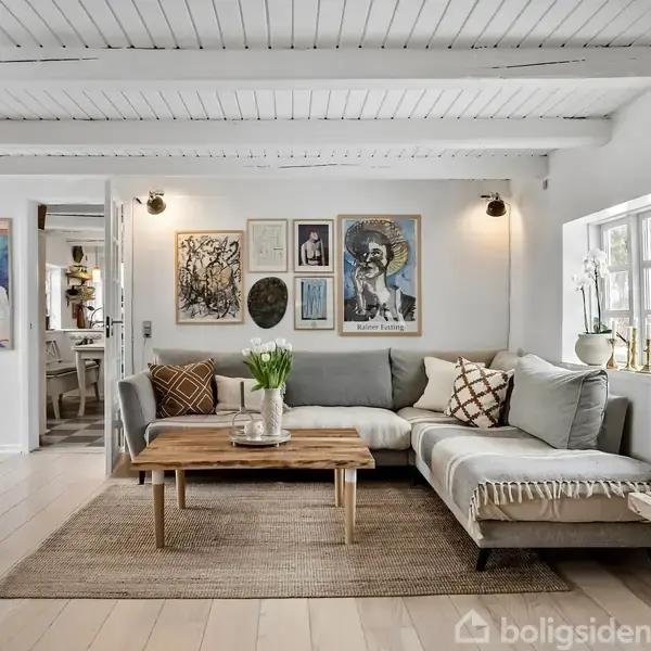 Stue med lyse møbler, hvor en hjørnesofa står ved et træbord. Kunst hænger på væggen. Rummet har træloft og trægulve, der tilføjer en hyggelig atmosfære.