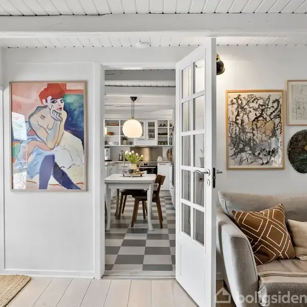 En lys stue med en åben dør viser et spiseområde. Malerier hænger på væggen over en beige sofa med puder. En stor lampe hænger over spisebordet.