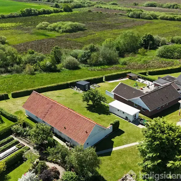 Huse med røde og mørke tage står stille i et grønt landskab, omgivet af velplejede haver og åbne marker.