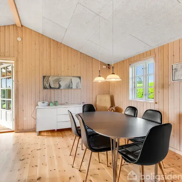 Rundt bord med seks sorte stole står i en lys spisestue med træbeklædning. To lamper hænger over bordet, og der er kunst på væggen. En dør åbner til udestue.