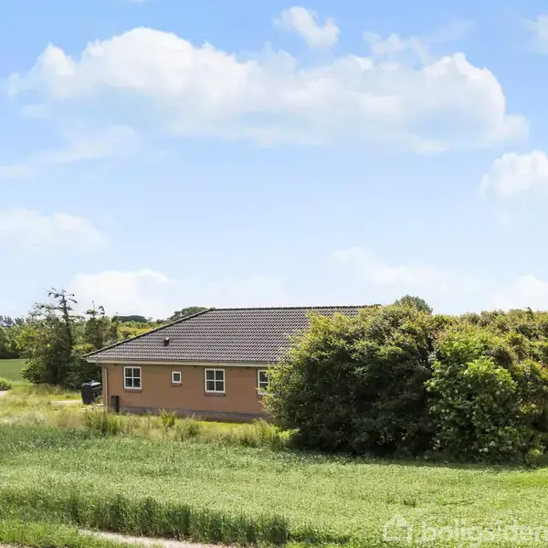 Et enetages hus står stille omgivet af grønne marker og buske, under en blå himmel med spredte skyer.