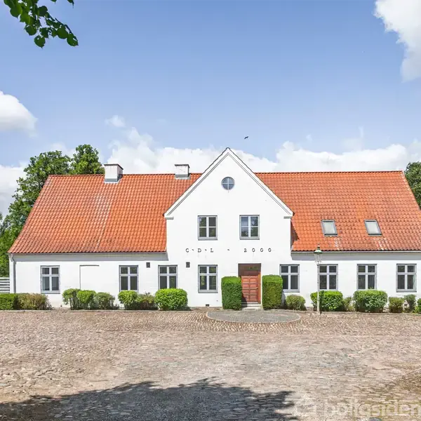 Hvidt hus med rødt tegltag står foran en grusplads, omgivet af grønne buske og træer. På bygningens front står "Opført 1866". Klar blå himmel og skyer i baggrunden.