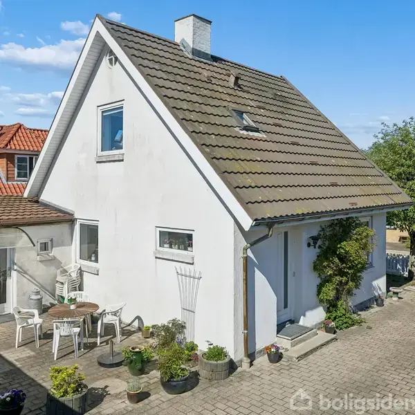 Et hvidt hus med skråt tag og karnap. En mindre terrasse foran med urtepotter og havemøbler. Omgivet af asfalt og andre huse under en klar blå himmel.