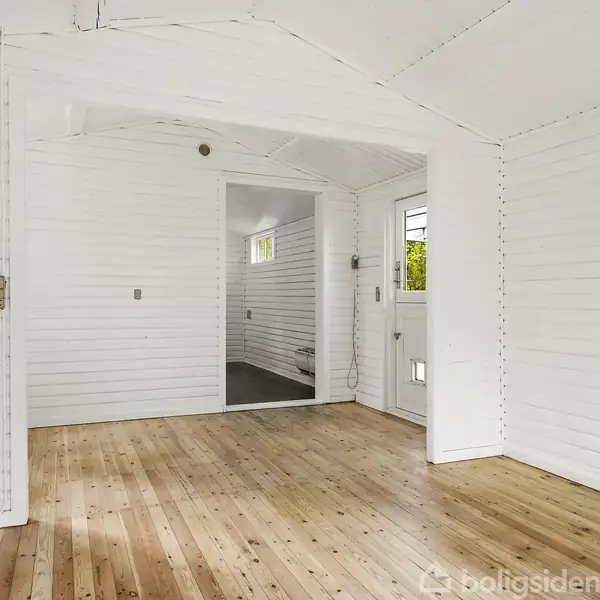 Et lyst rum med hvide trævægge og trægulve. En åben dør fører til et lille, tilstødende rum. Små vinduer giver naturligt lys. Ingen tekst fundet.