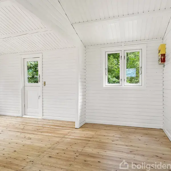 Et lyst rum med hvide vægge og træloft står tomt. To vinduer og en dør i rummet giver udsigt til grønne omgivelser. En brandslukker hænger på væggen.