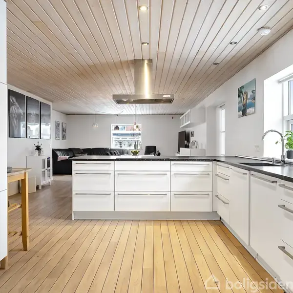 Køkken med hvide skabe og en sort bordplade, der åbner ind til en stue med sofa og billeder på væggen. Trægulv og lyse træpaneler i loftet.