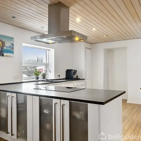 Et moderne køkken med en stor køkkenø med sort bordplade. Der hænger en kogeplade og emhætte over øen. Maleri og vindue i baggrunden. Trægulv og lofter.