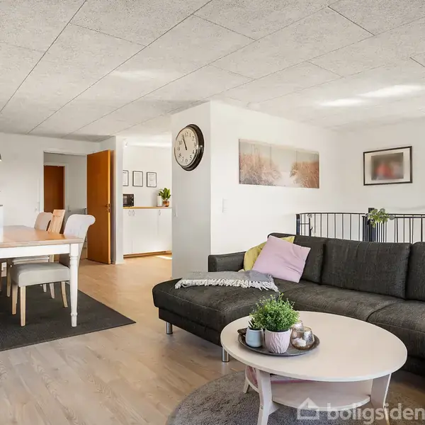 Sort sofa står i en lys stue med et rundt sofabord foran. Til venstre et spisebord med seks stole. Vægge har kunst og en stor ur.