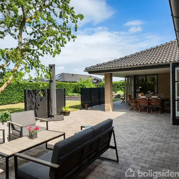 Terrasse med siddeområde og sofa, omgivet af frodig grøn beplantning, under blå himmel i et moderne husmiljø. Spisearrangement under overdækket sektion på højre side.