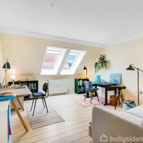 To skriveborde står i et lyst arbejdsrum med skråvinduer. Begge skriveborde har bøger og lamper. En sofa og en plante hænger ved vinduet, med lyse træplanker på gulvet.