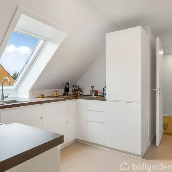 Et lyst køkken med hvide skabe og et stort ovenlysvindue, som giver naturligt lys. Køkkenredskaber står langs væggen. Til højre fører en åben dør til en korridor.